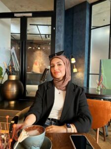 Nahl is wearing a white shirt and black blazer, with a brown headscarf and sunglasses.